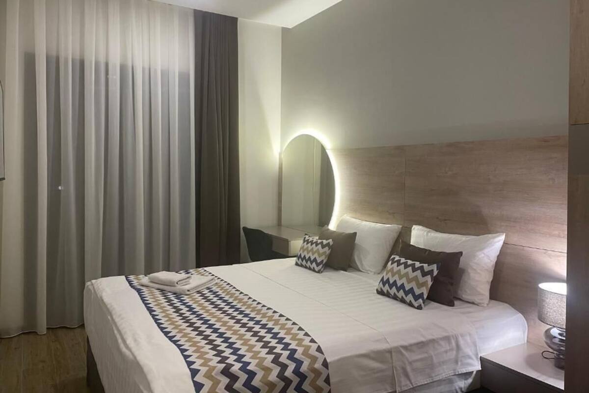 Moderni hotellihuone, jossa on suuri sänky, yöpöytä, seinään kiinnitetty peili ja ikkuna, jossa on verhot.
