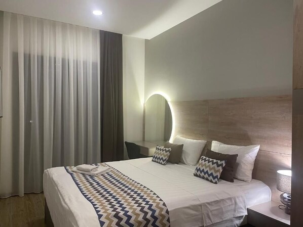 Moderni hotellihuone, jossa on suuri sänky, yöpöytä, seinään kiinnitetty peili ja ikkuna, jossa on verhot.
