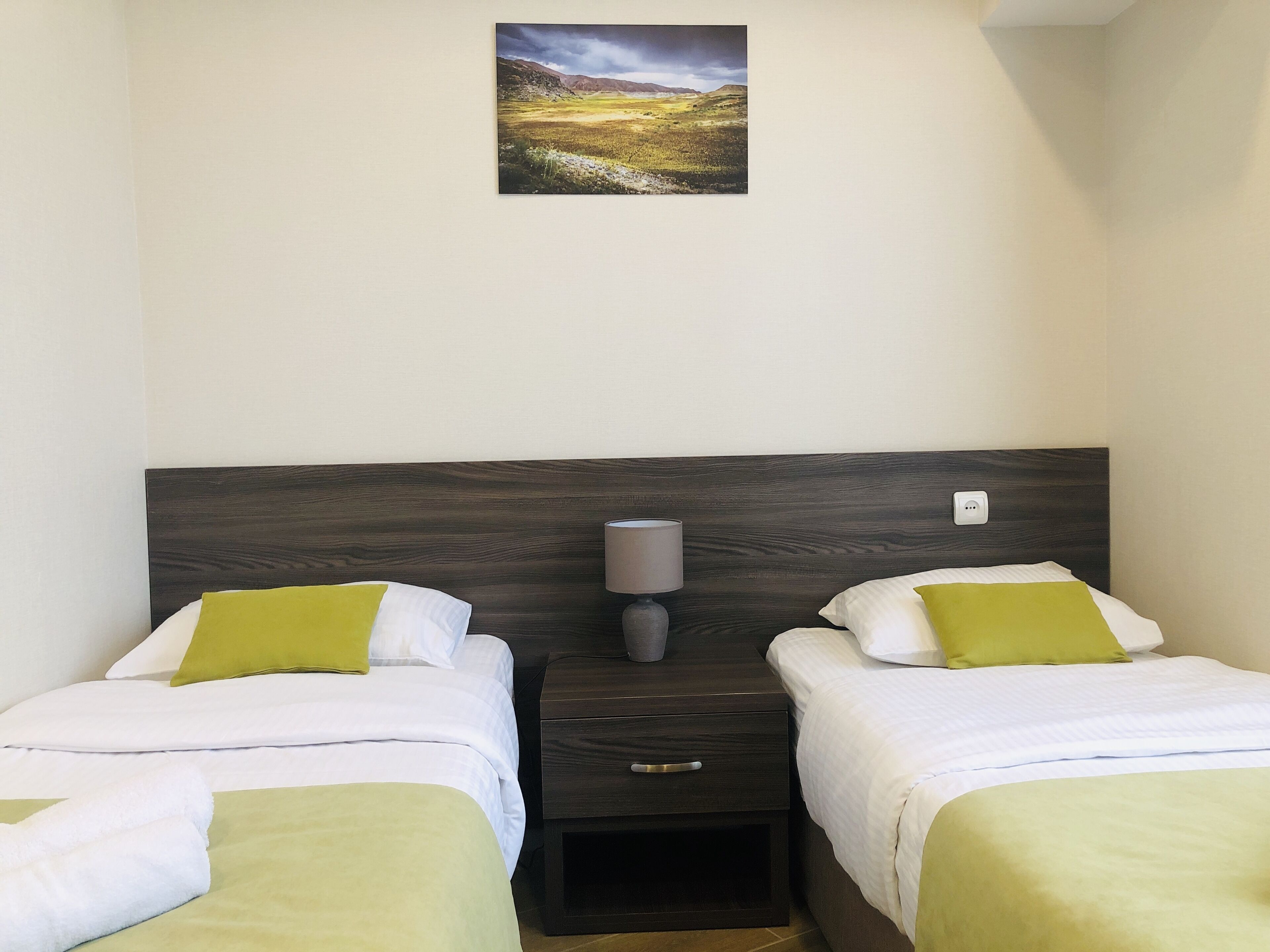 A hotel room with two beds, each with white bedding and green pillows, a wooden headboard, a bedside table with a lamp, and a painting of a landscape on the wall.