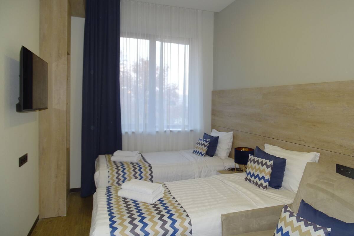 Hotellihuone, jossa on kaksi sänkyä, puinen pääty, seinälle asennettu televisio ja ikkuna, jossa on verhot.