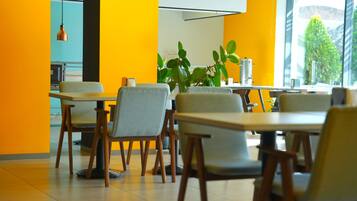 A modern dining area with wooden tables and chairs, a large window with a view of greenery, and a bright yellow accent wall.