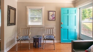 Una habitación con dos sillas, una mesita, una ventana con persianas, una puerta azul y un cuadro en la pared.