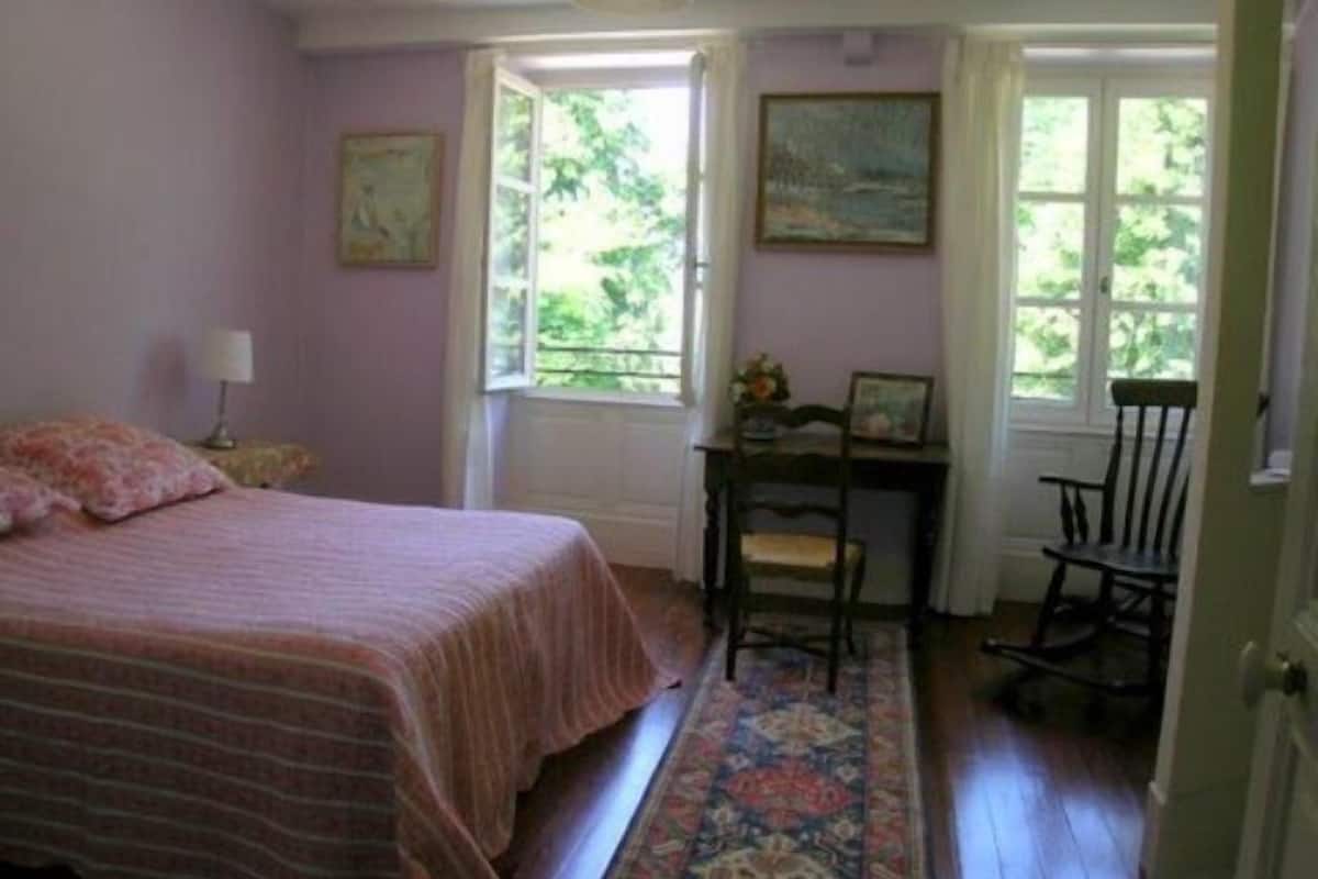 A bedroom with a bed, a desk, and two chairs. There are windows with views of greenery outside.