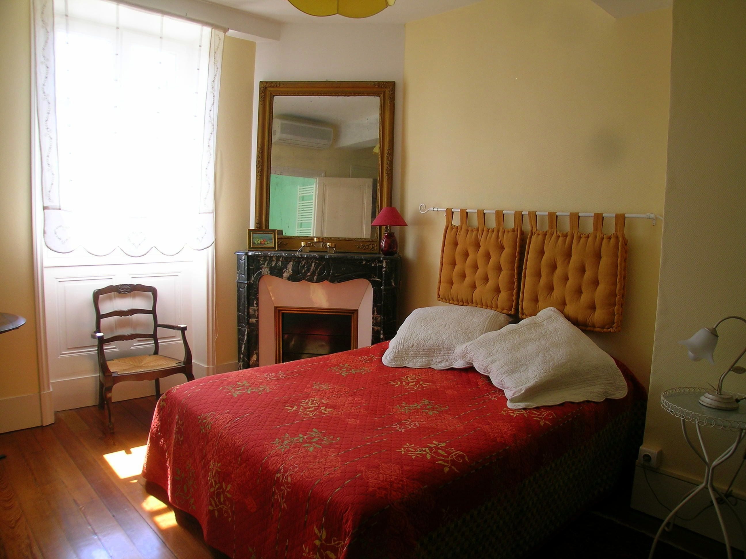 A bedroom with a bed, a chair, a fireplace, a mirror, and a lamp.