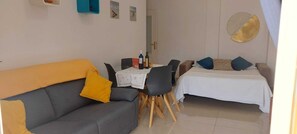 Un espace de vie compact comprenant un canapé gris, une petite table à manger et un lit recouvert d’une courtepointe blanche.