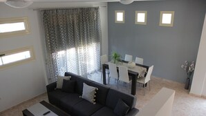 A living room with a dark grey sofa, a dining table with chairs, and a window with patterned curtains. - Platoni Elite Villas (Rhodes)
