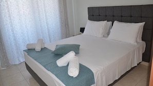 A neatly made bed with white linens and a teal throw, a grey upholstered headboard, and a white curtain with a subtle pattern. - Platoni Elite Villas (Rhodes)