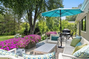 Relax in a serene outdoor space with a wicker sofa, dining table, and barbecue grill, surrounded by lush greenery and a wooden deck.