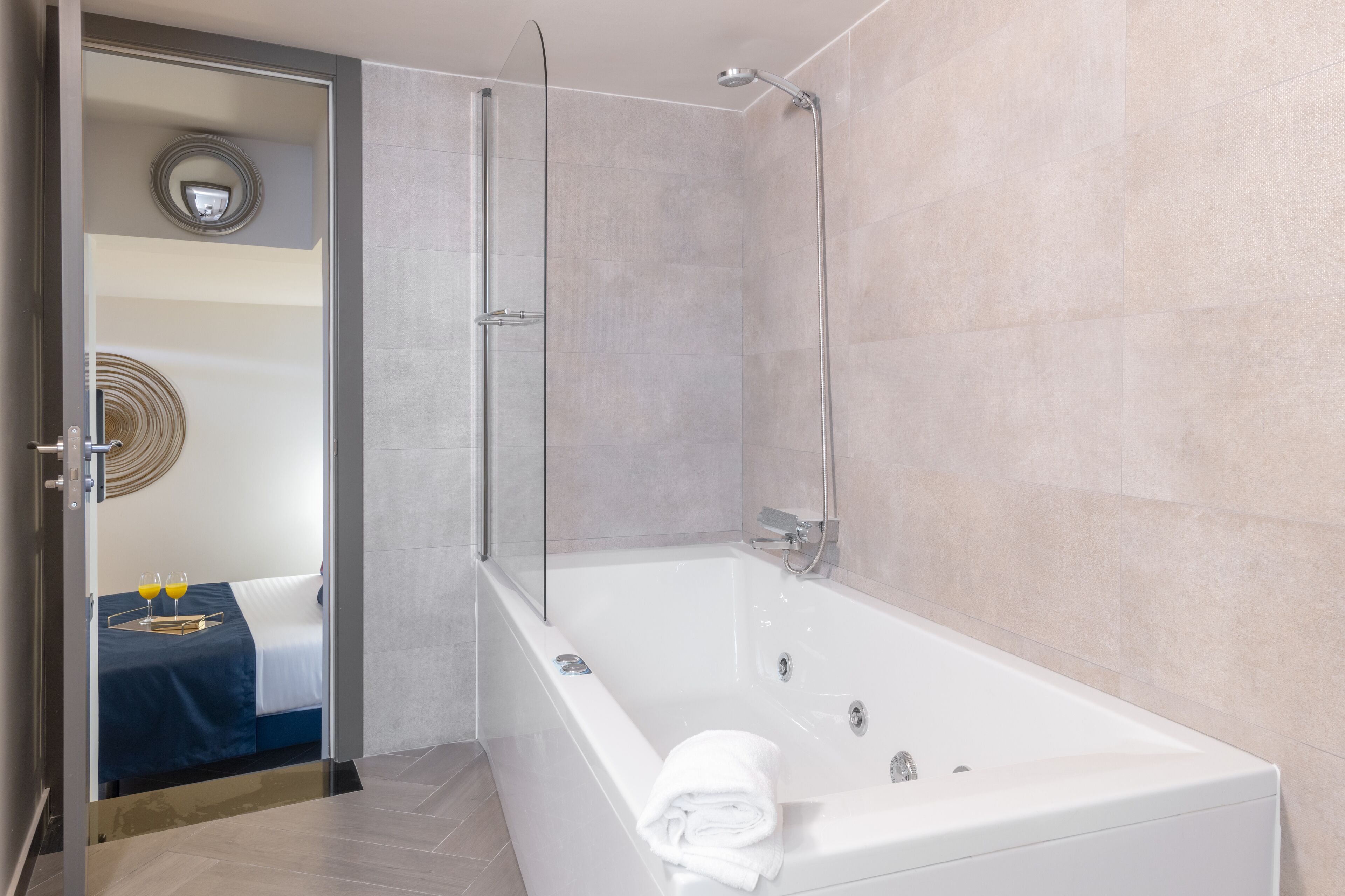 Une salle de bain moderne avec une douche à parois de verre, une baignoire blanche et un lit visible, recouvert d’une couverture bleue, avec deux verres de jus d’orange posés à proximité.