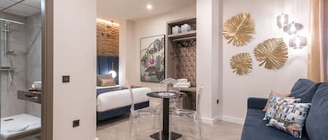 Une chambre d’hôtel moderne comprenant un lit, un coin salon avec une table et des chaises, un espace douche et des œuvres d’art décoratives au mur.