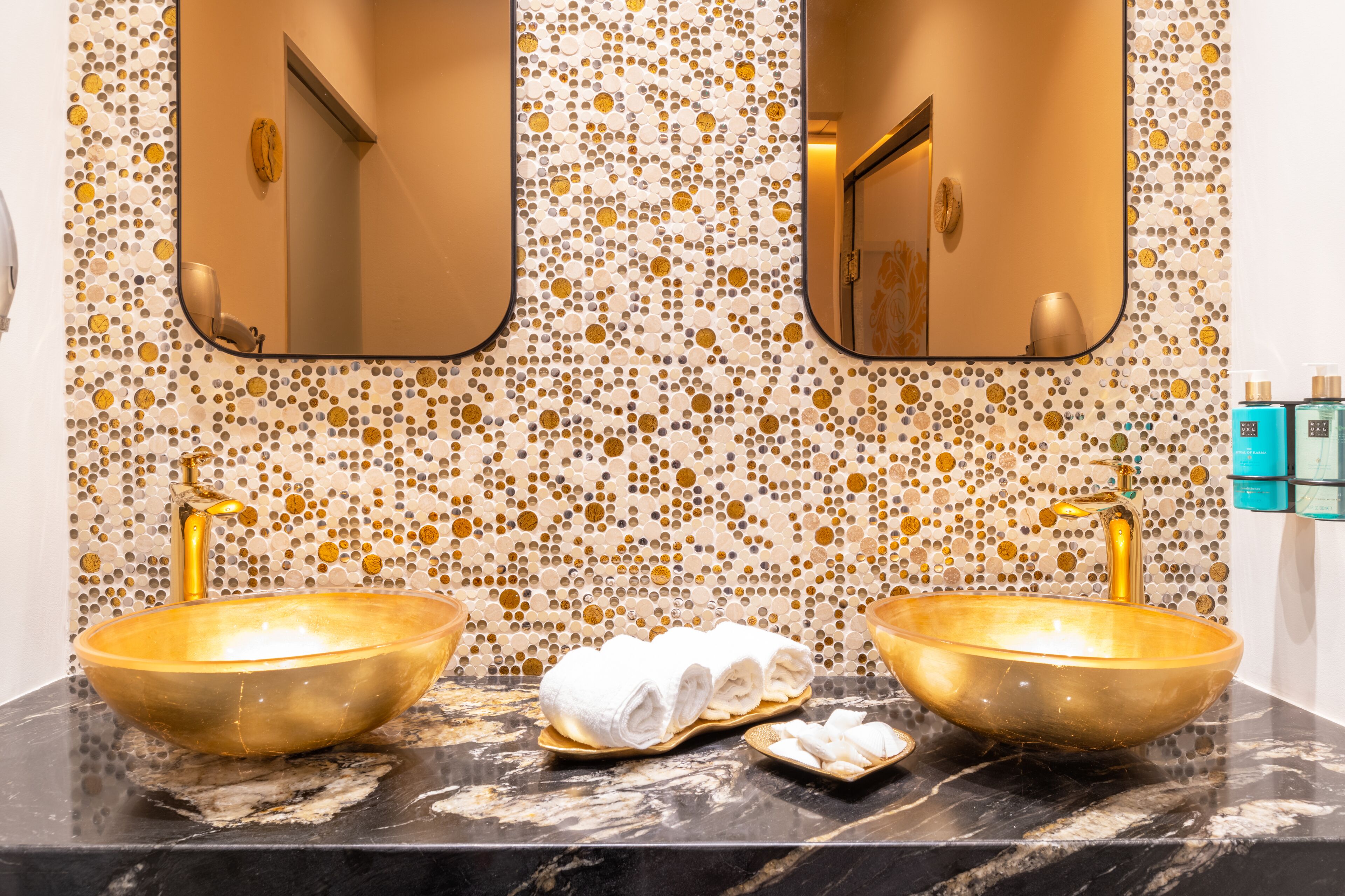 A bathroom with two gold sinks, a marble countertop, and a mosaic tile wall.