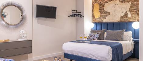 A modern bedroom with a brick accent wall, a flat-screen TV, a bedside table with decorative items, and a blue upholstered bed.