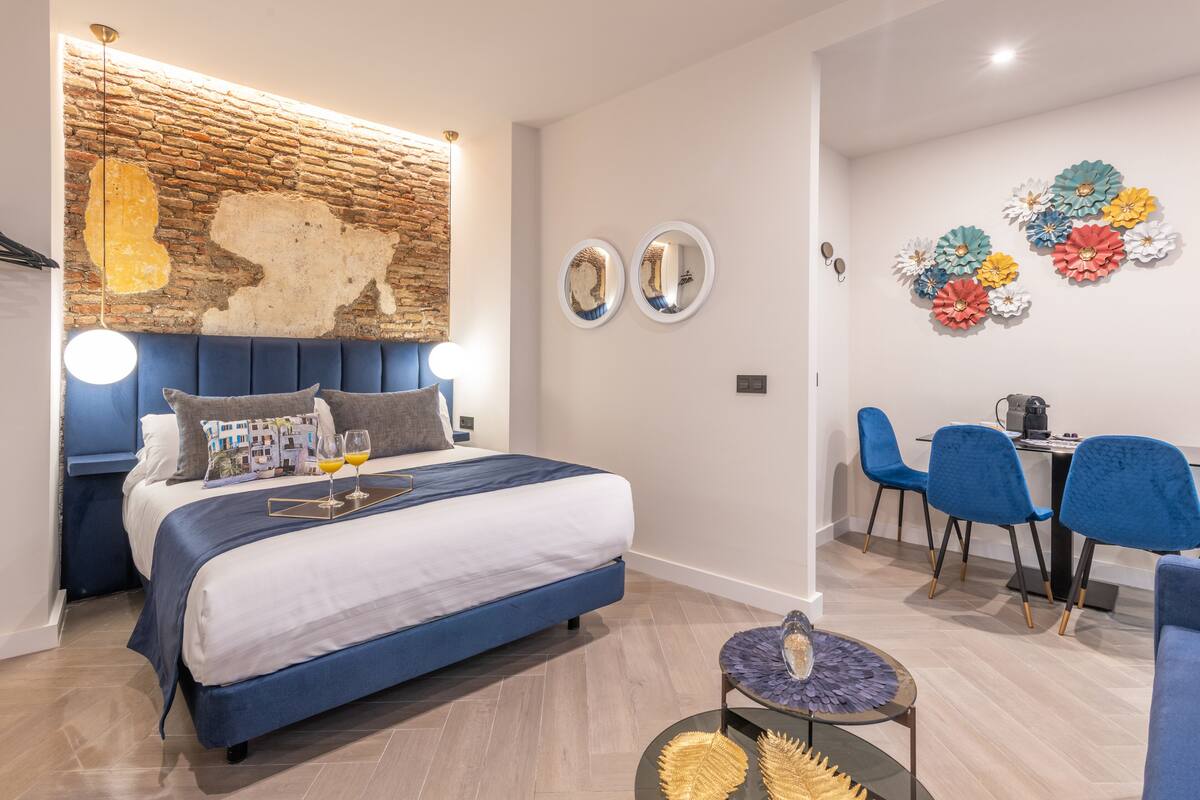 A modern bedroom with a brick accent wall, a large bed with a blue headboard, two round mirrors, and a dining area with blue chairs and a table.