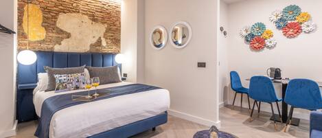 A modern bedroom with a brick accent wall, a large bed with a blue headboard, two round mirrors, and a dining area with blue chairs and a table.