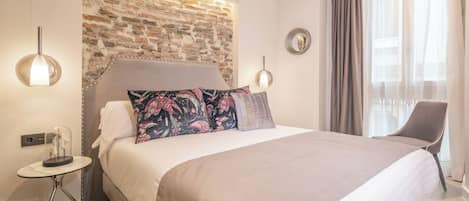 A modern hotel room with a brick accent wall, a bed with patterned pillows, a side table with a lamp, and a chair by the window.