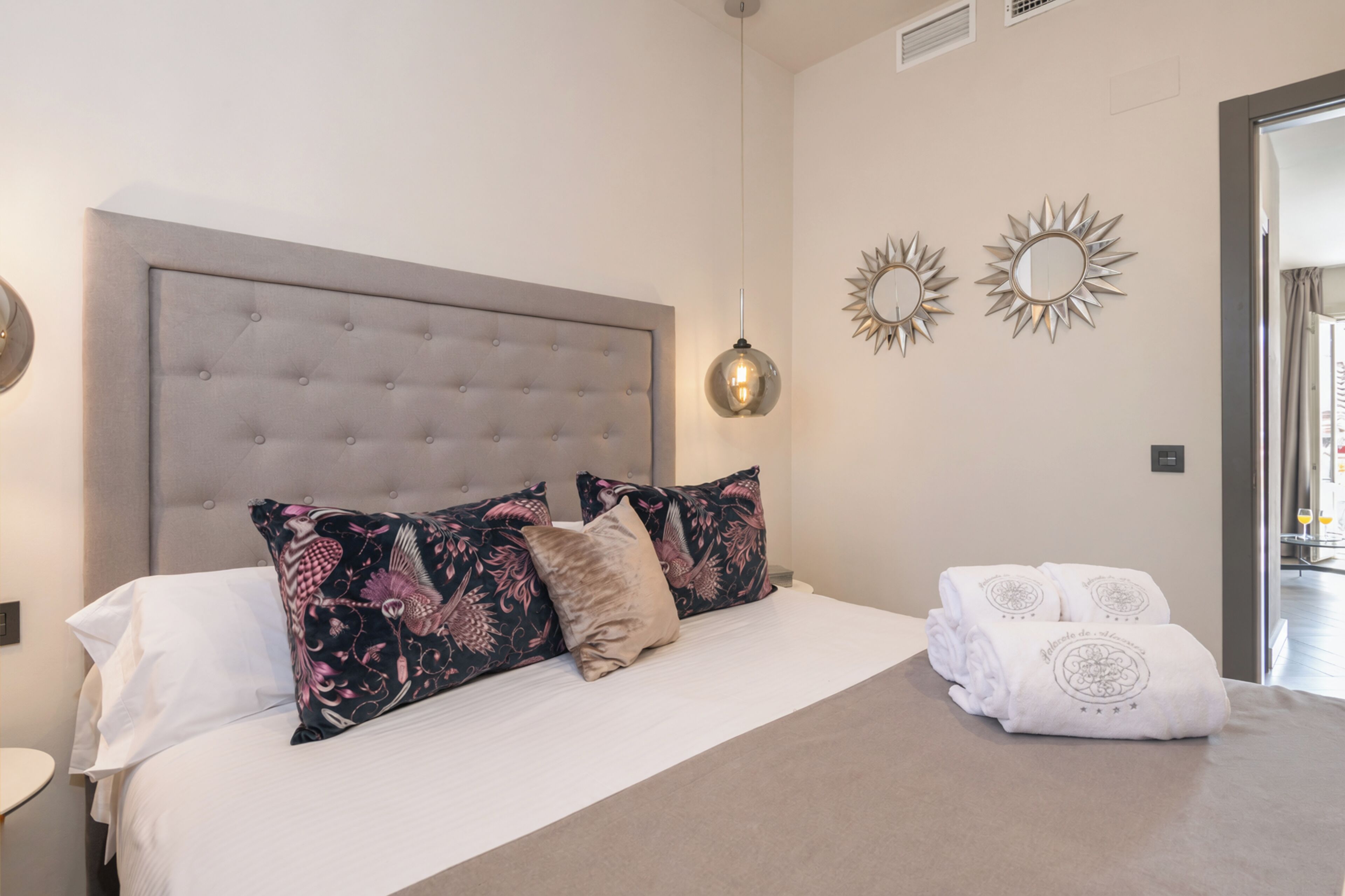 A bedroom with a tufted headboard, a hanging pendant light, decorative wall mirrors, and neatly folded towels.