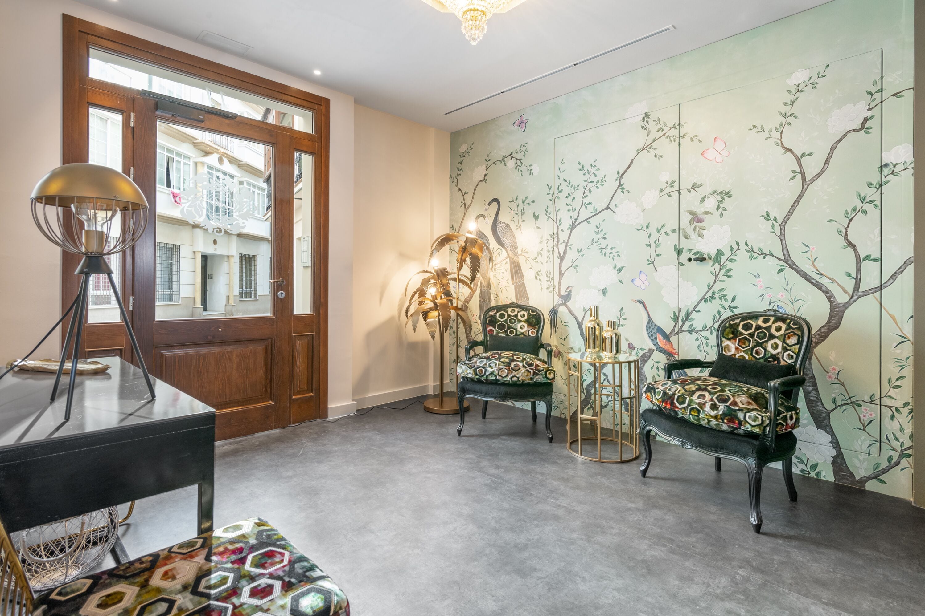 A room with a wooden door, a table with a lamp, two patterned chairs, and a wall with a tree mural.