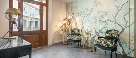 A room with a wooden door, a table with a lamp, two patterned chairs, and a wall with a tree mural.