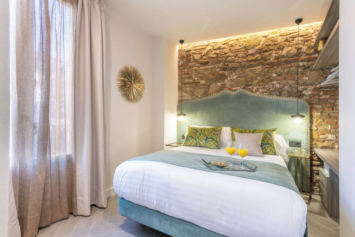 A bedroom with a stone wall, a bed with a green headboard, a bedside table with a glass of orange juice, and a curtain on the window.