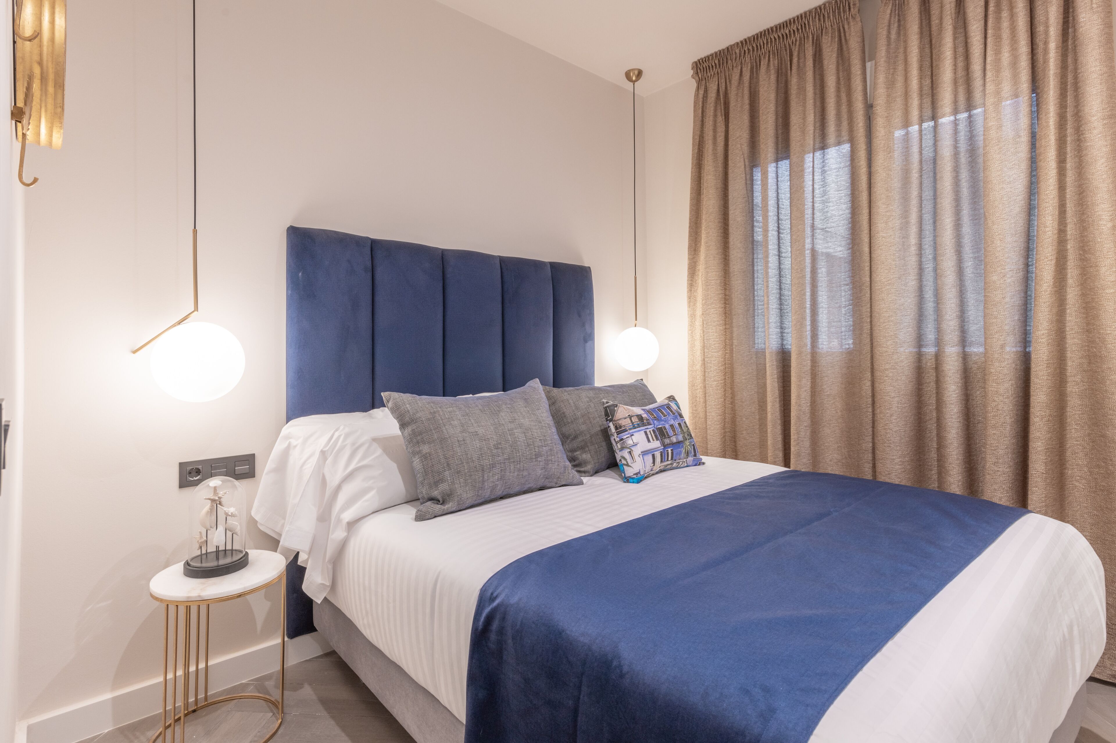 Una cama bien hecha con un cabecero azul, una mesita de noche con una lámpara y una ventana con cortinas.