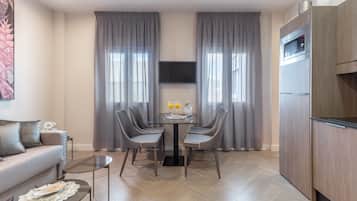 A modern living room with a sofa, a glass coffee table, and a dining table with chairs. There is a flat-screen TV mounted on the wall, a built-in oven, and a refrigerator.