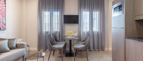 A modern living room with a sofa, a glass coffee table, and a dining table with chairs. There is a flat-screen TV mounted on the wall, a built-in oven, and a refrigerator.