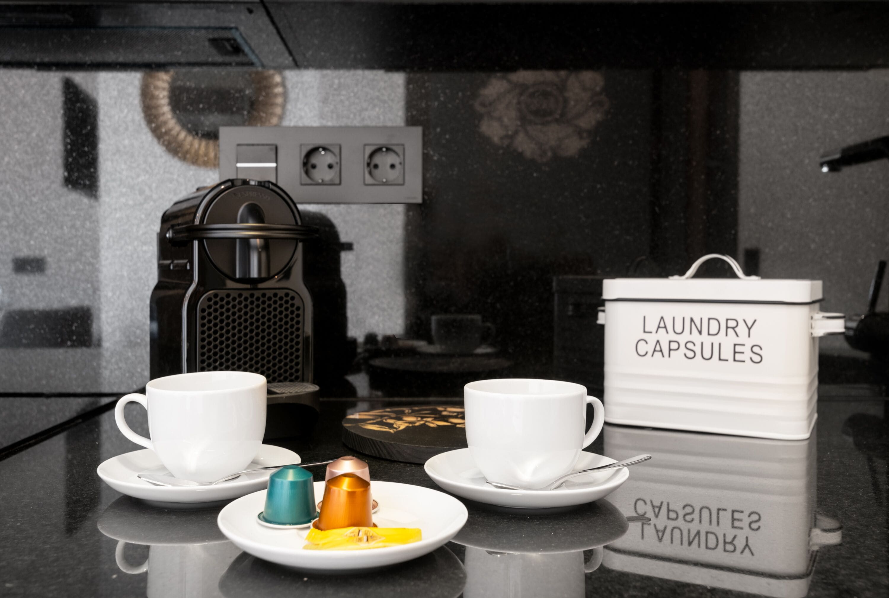 A kitchen counter with two white cups, a coffee maker, and a container labeled "laundry capsules".