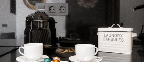 Una encimera de cocina con dos tazas blancas, una cafetera y un recipiente con cápsulas para la lavadora.