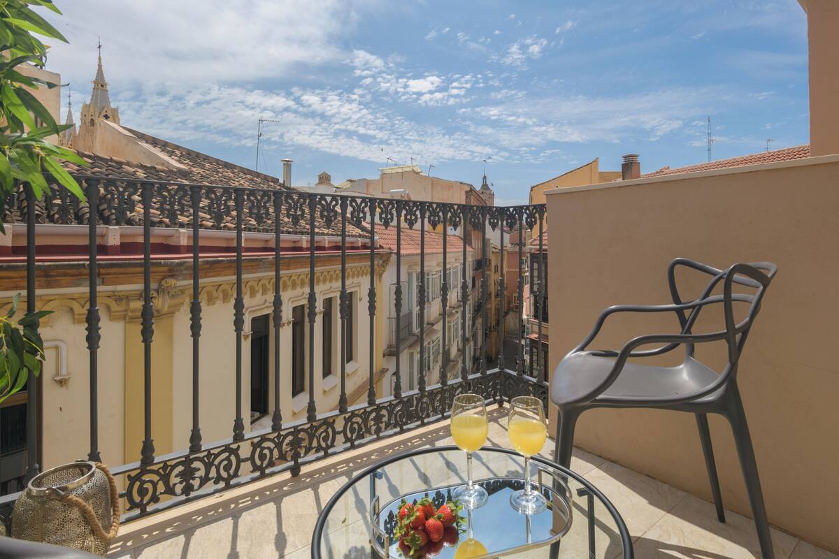 A balcony with a glass table, two glasses of orange juice, a bowl of strawberries, and a chair.