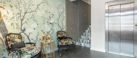 A modern lobby with two upholstered chairs, a decorative wall mural, a marble staircase, and a stainless steel elevator.