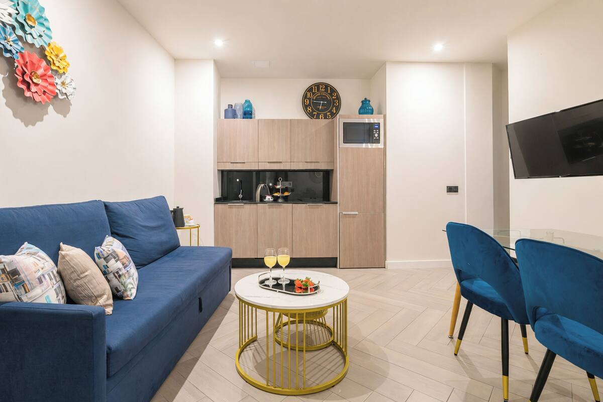 A modern living room with a blue sofa, a round gold table, and a kitchen in the background.