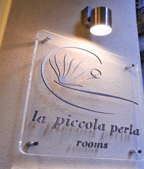 Une plaque en verre transparent, ornée d’un motif stylisé représentant un escargot, et portant l’inscription « la piccola perla rooms », indiquant une chambre ou un espace spécifique à l’intérieur d’un hôtel.