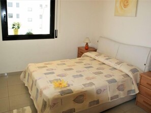 A bedroom with a bed, a nightstand, a lamp, a window with a view of buildings, and a wall with a painting.