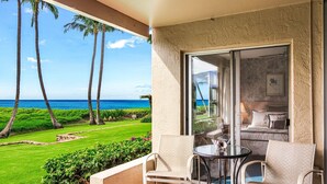 A balcony with a view of the ocean and palm trees, a glass door leading to a bedroom with a bed and a nightstand, and a small table with two chairs. - Menehune 122 Beautiful Ocean Views in Beautiful Condo Home (Kihei)
