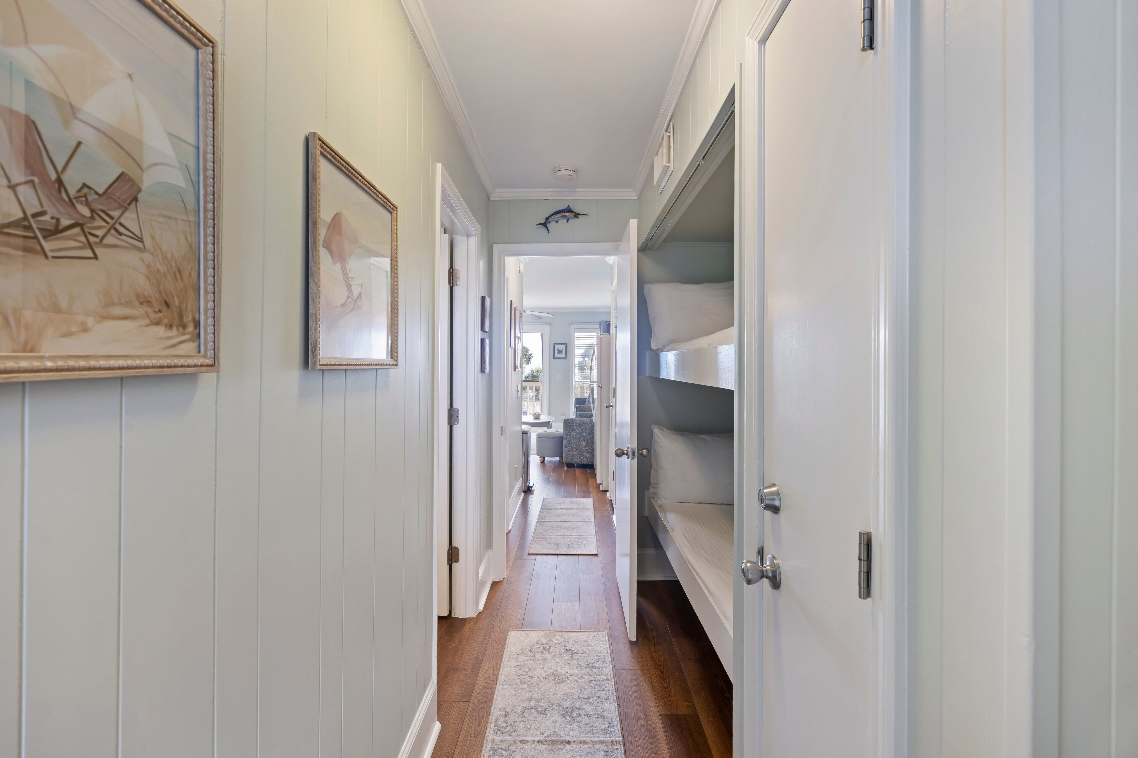 A hallway with wooden floors, white walls, and a door leading to a bedroom. There are framed pictures on the walls and a rug on the floor.