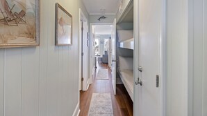 Un couloir avec un plancher en bois, des murs blancs et une porte donnant sur une chambre. Des tableaux encadrés ornent les murs et un tapis recouvre le sol.