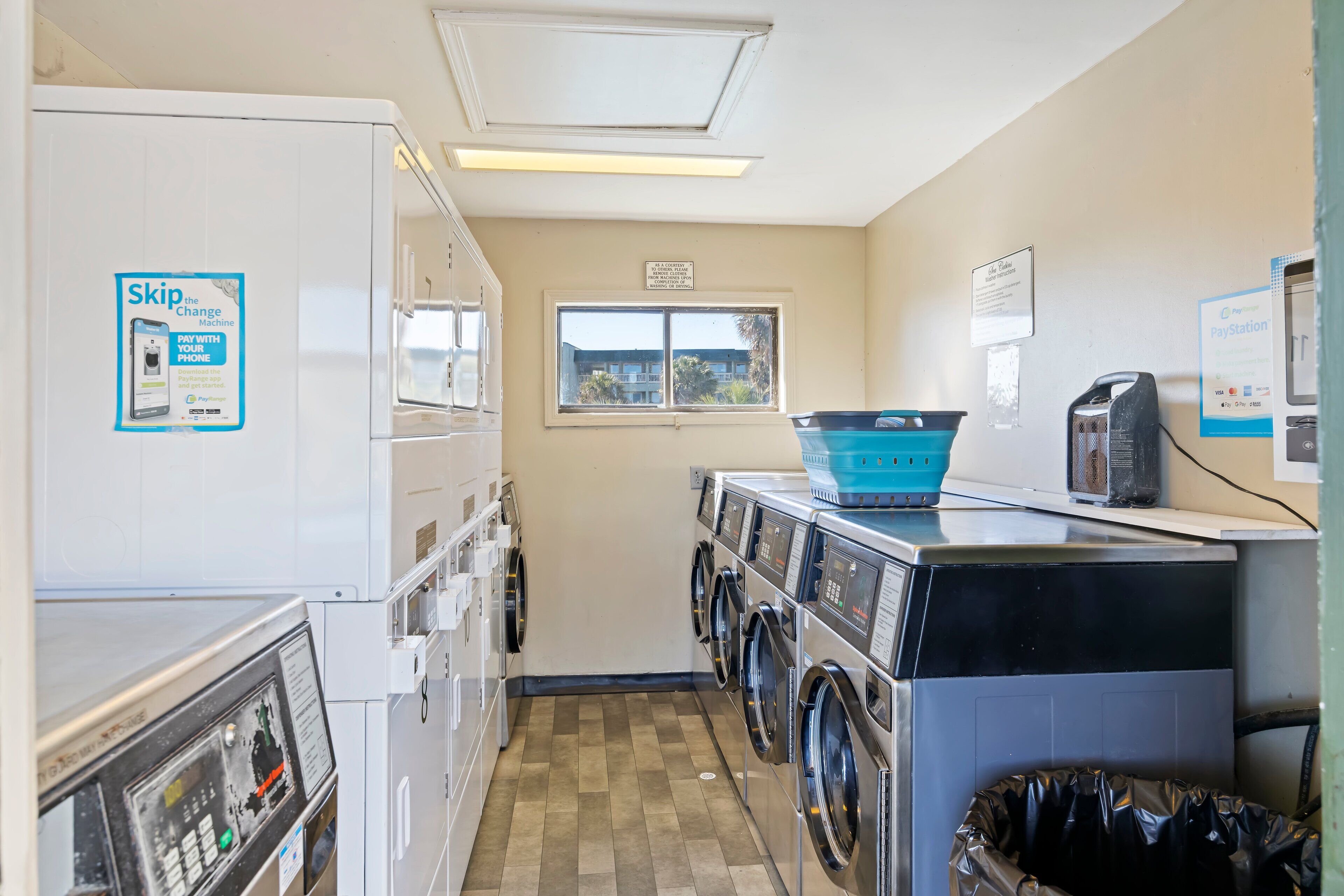 A laundry room with a row of washers and dryers, a blue basket on the dryer, a trash can, and a sign advertising a Pay with Phone service.