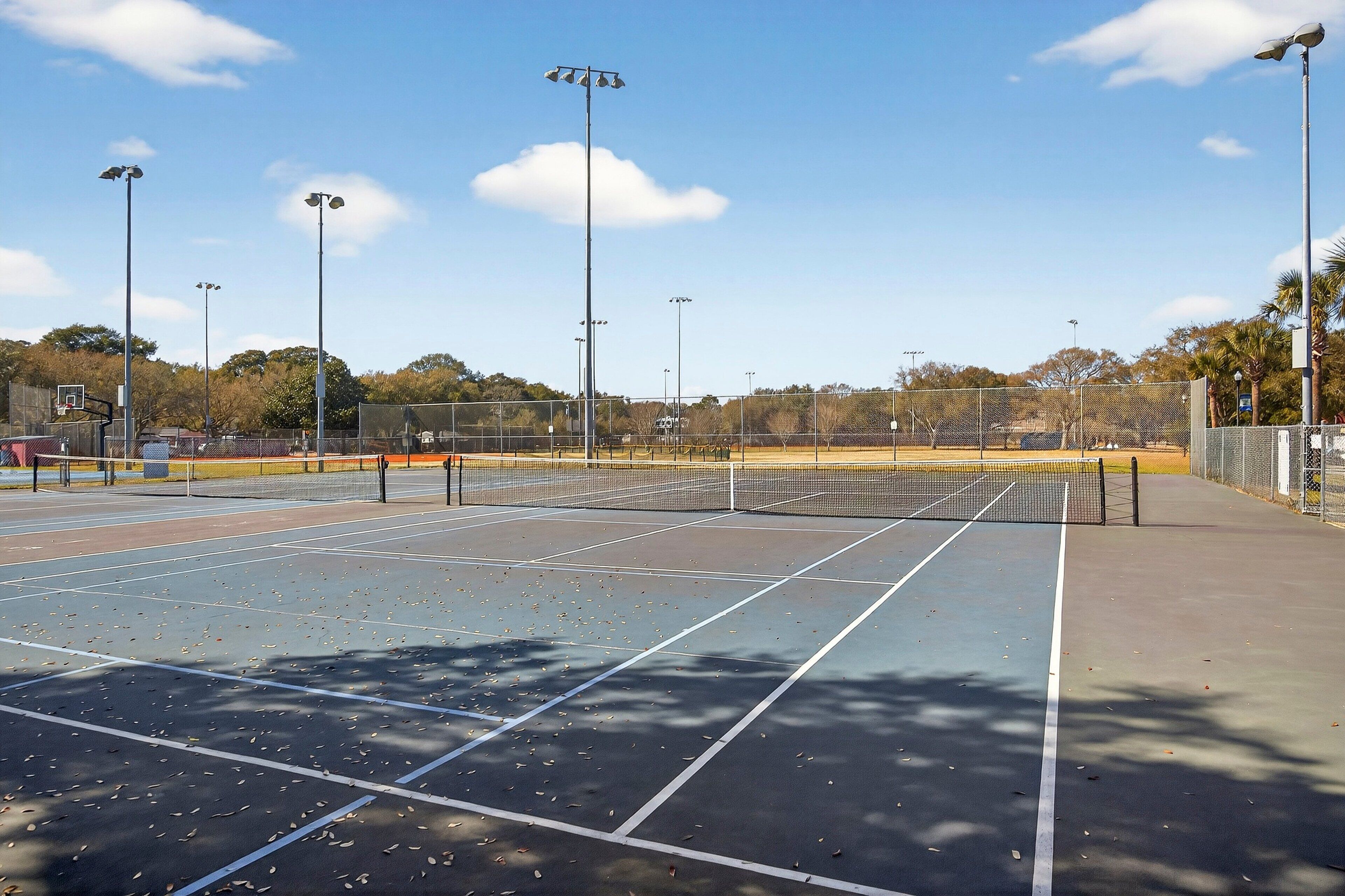 An outdoor tennis court with a net, surrounded by a fence, and street lamps.