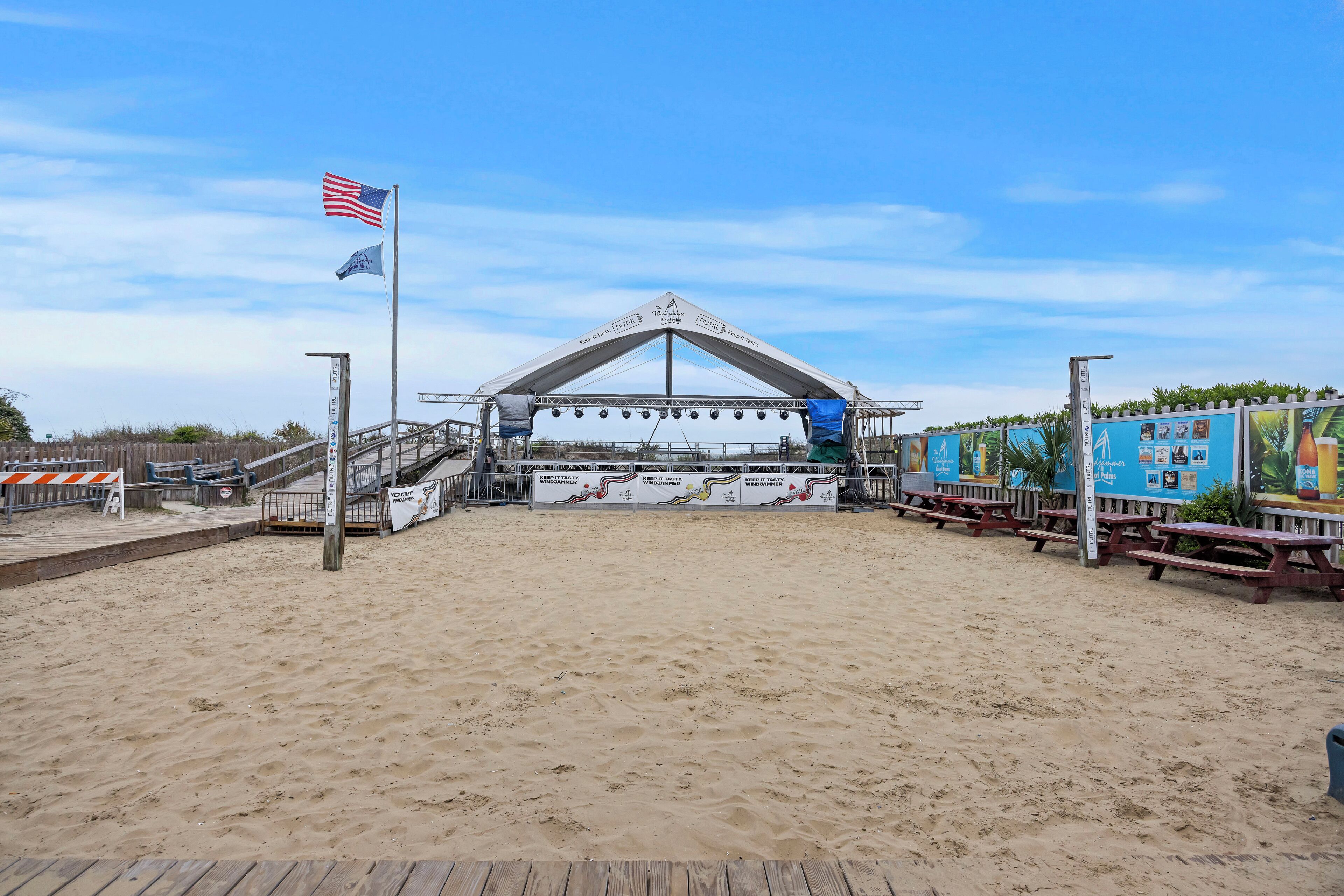A beachside stage with a tent, a flag, and a wooden walkway leading to it.