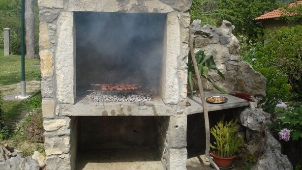 Un four extérieur en pierre, dans lequel brûle un feu, entouré de verdure et bordé d’un mur de pierre rustique.