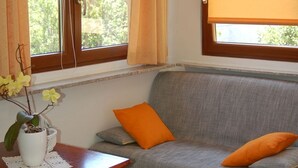 Une pièce chaleureuse avec un canapé gris, des coussins orange, une petite table en bois ornée d’un vase de fleurs et une fenêtre avec un rideau.
