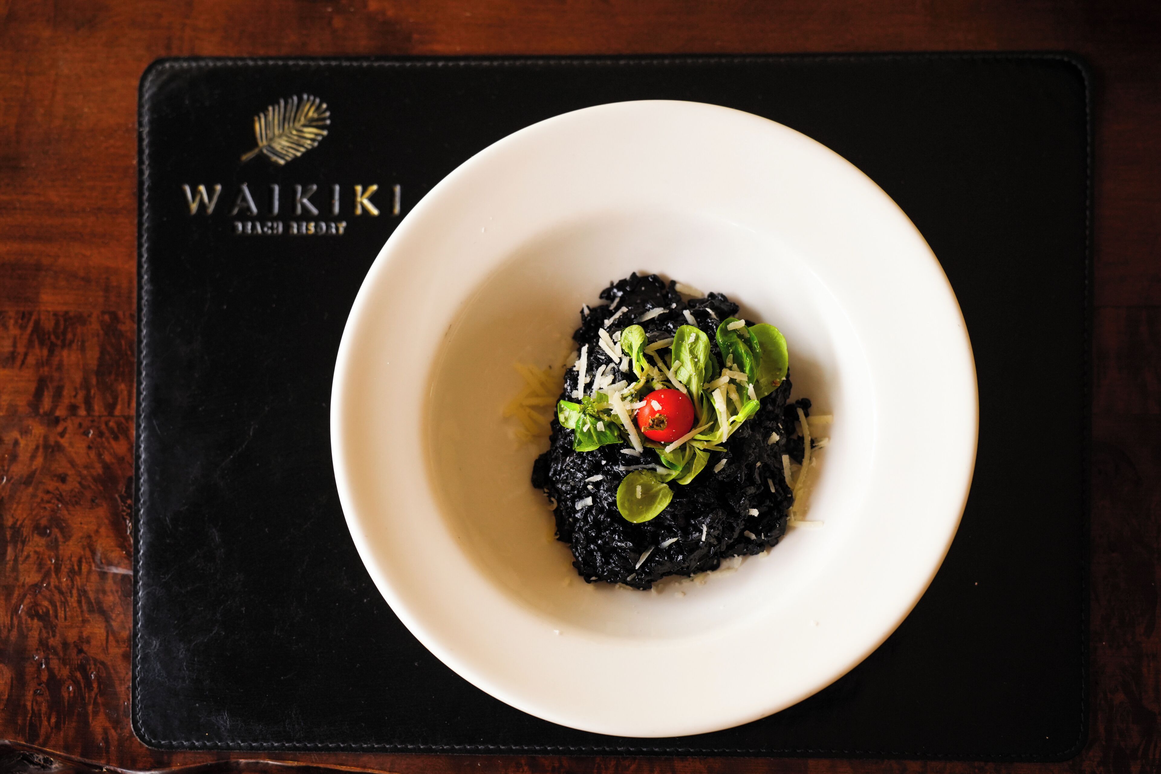 Un tazón blanco con arroz negro, verduras y una cereza, sobre un camino de mesa negro con el texto 'WAIKIKI BEACH RESORT'.