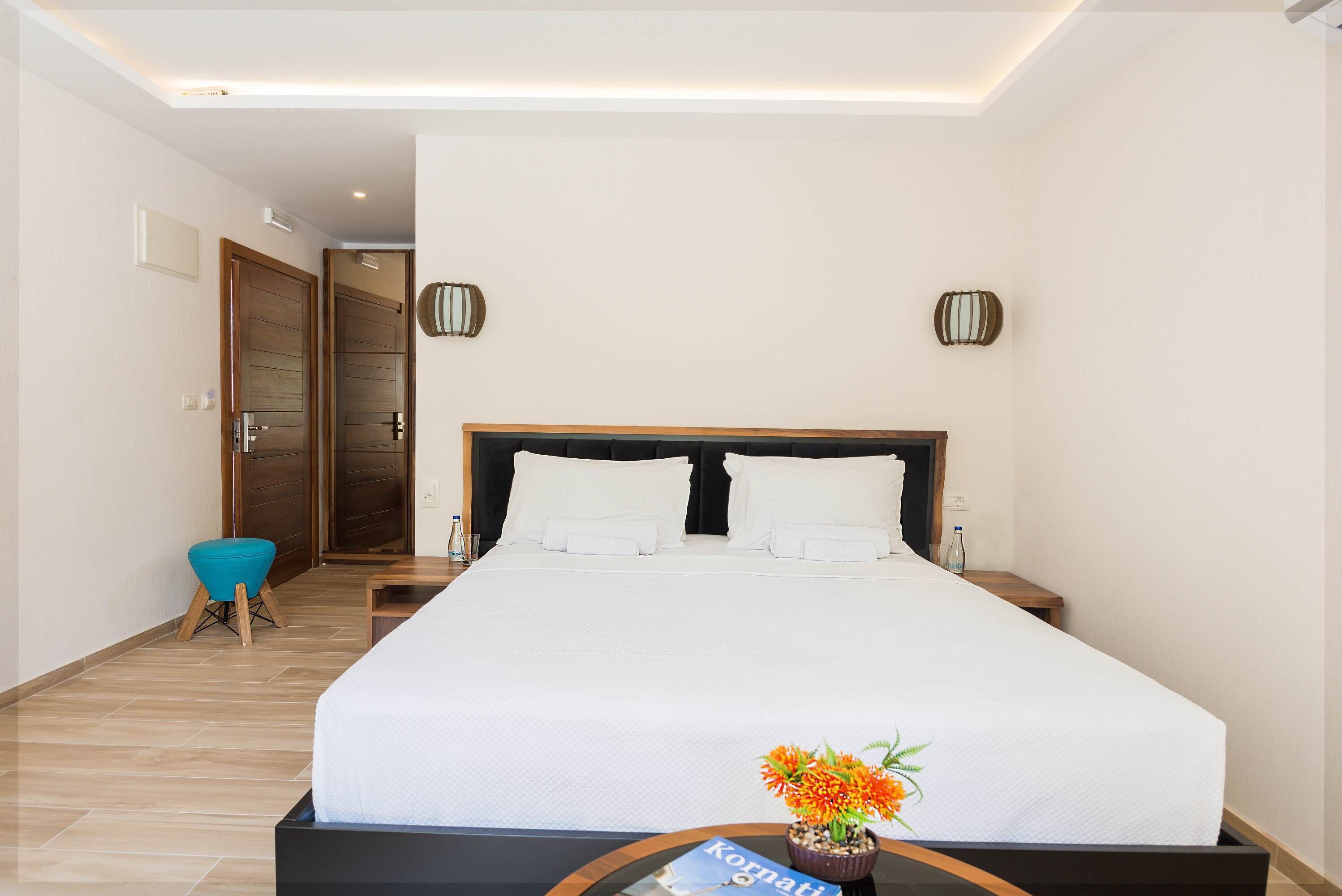 Una camera d'albergo con un letto grande, due comodini, una piccola poltrona blu e un tavolino con una bottiglia.