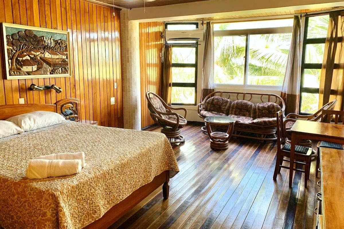 A room with a bed, a sofa, a dining table, and a chair. There is a window with curtains and a painting on the wall.
