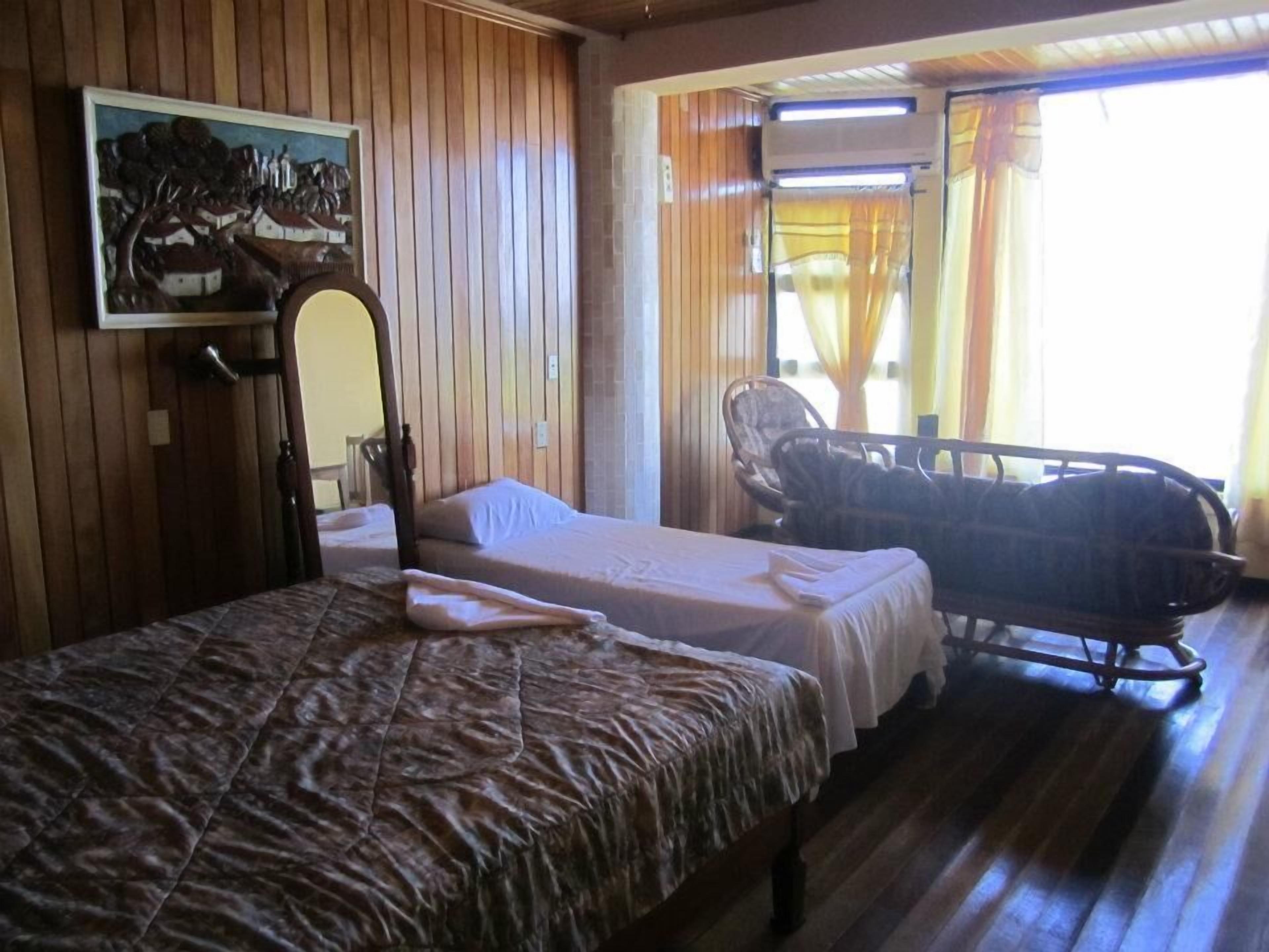 A room with two single beds, a wooden wall, a painting, a chair, and a window with curtains.