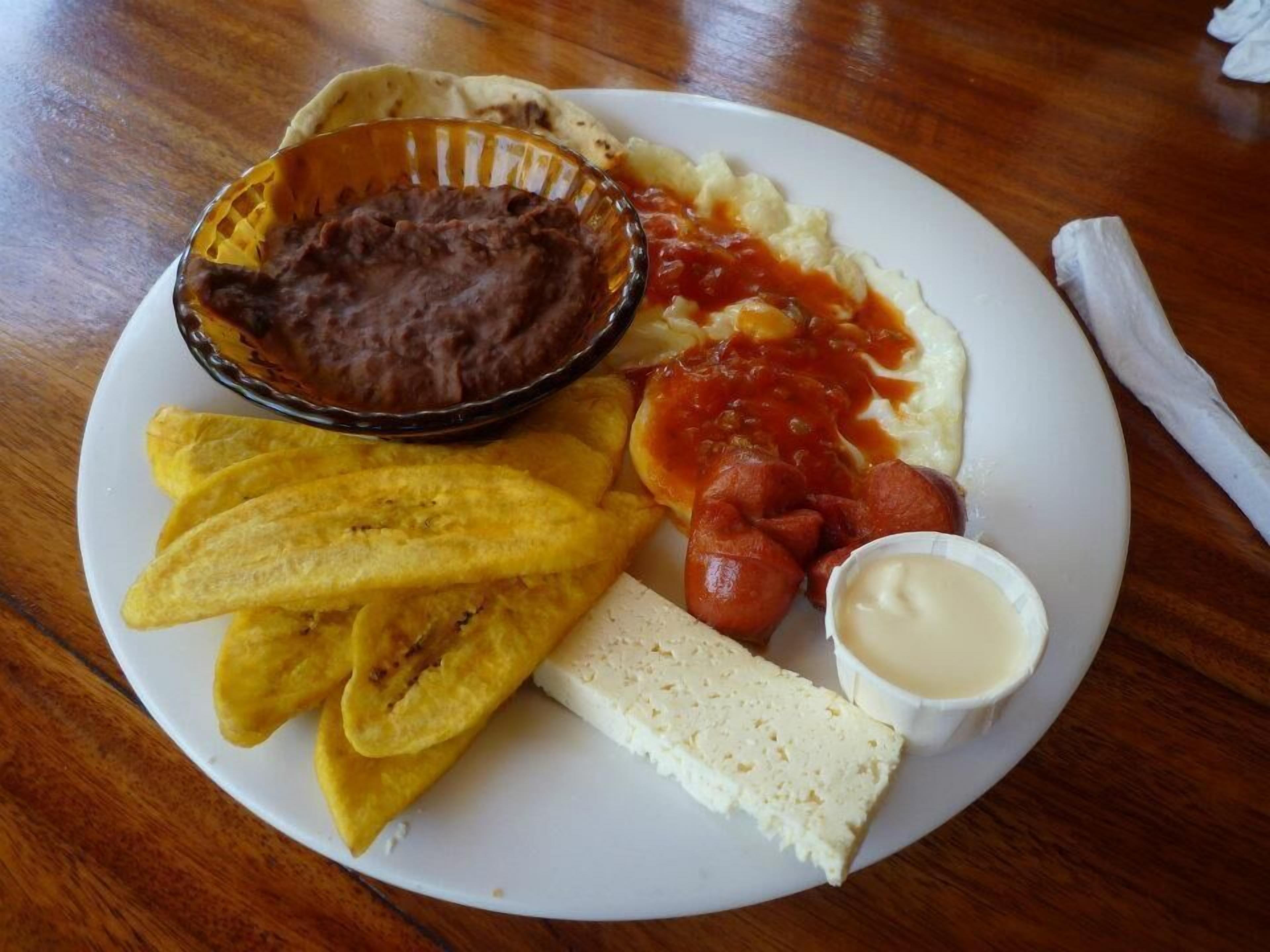 Un plato con una variedad de alimentos, incluyendo frijoles, salsa de tomate, plátanos fritos, un hot dog y un recipiente pequeño con salsa.