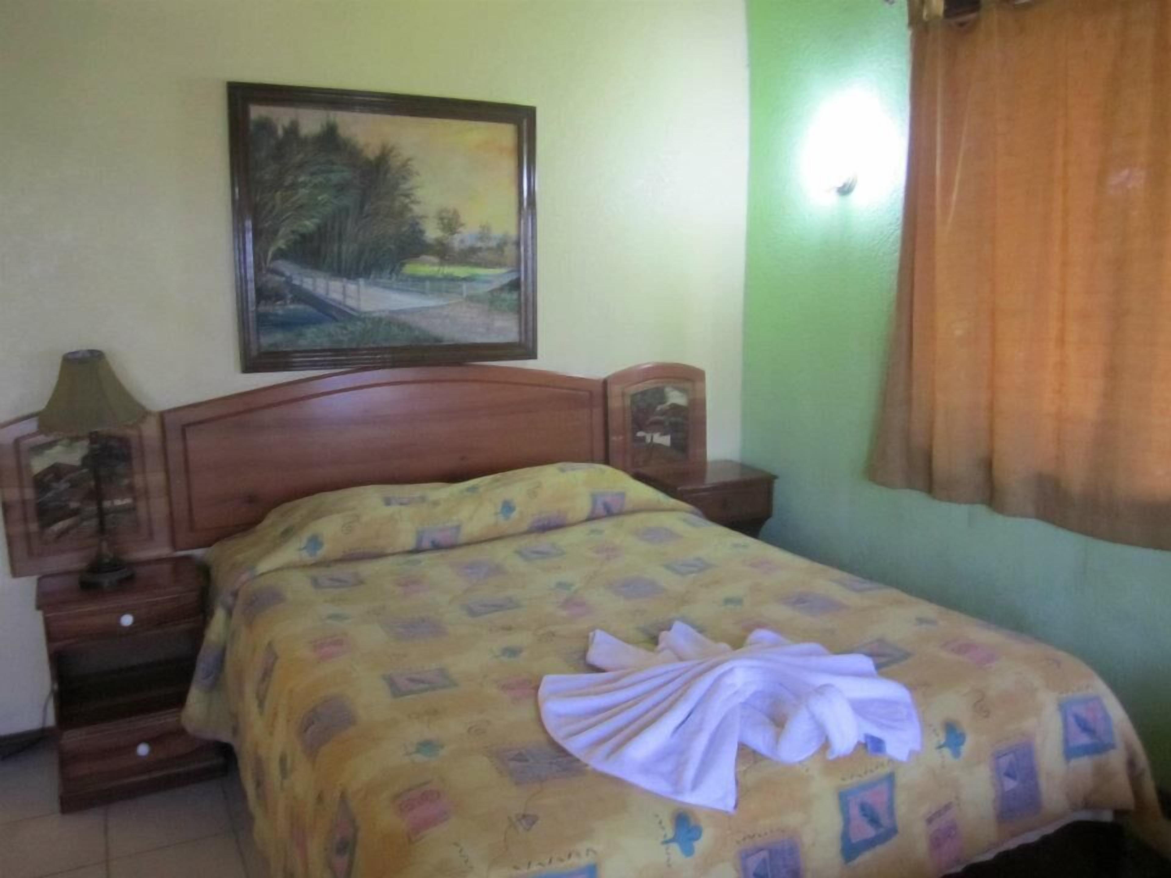 A bedroom with a bed, a nightstand, a lamp, a painting on the wall, and a window with curtains.