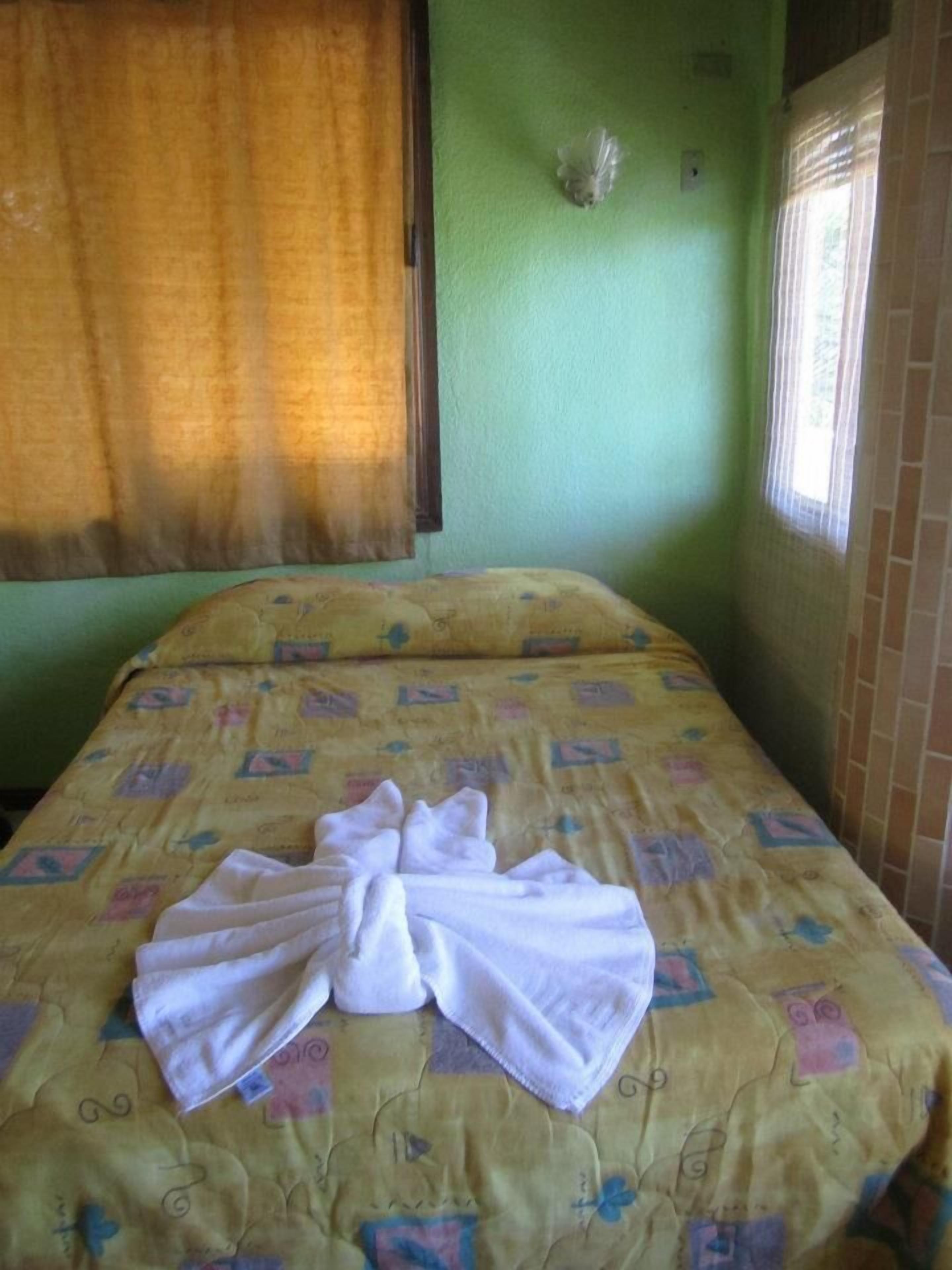 A room with a bed covered in a patterned bedspread, a folded white towel on the bed, and a window with curtains.