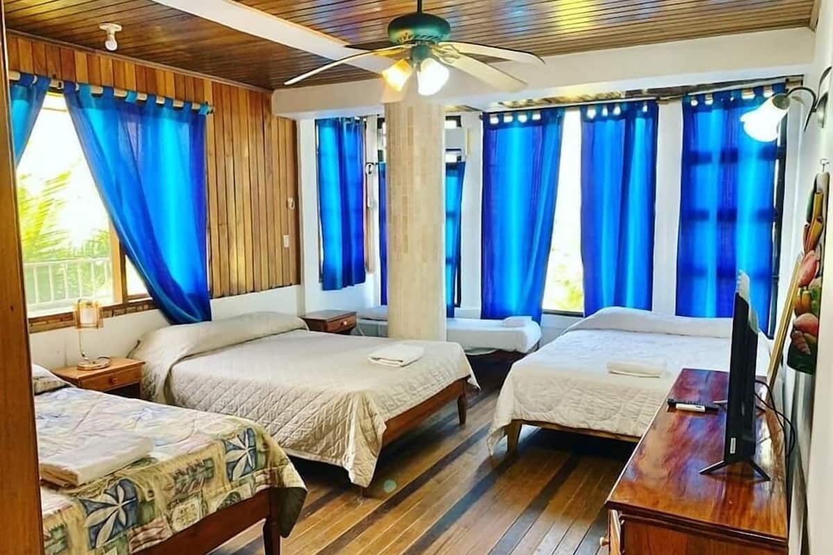 A room with two beds, wooden flooring, a ceiling fan, and large windows with blue curtains.