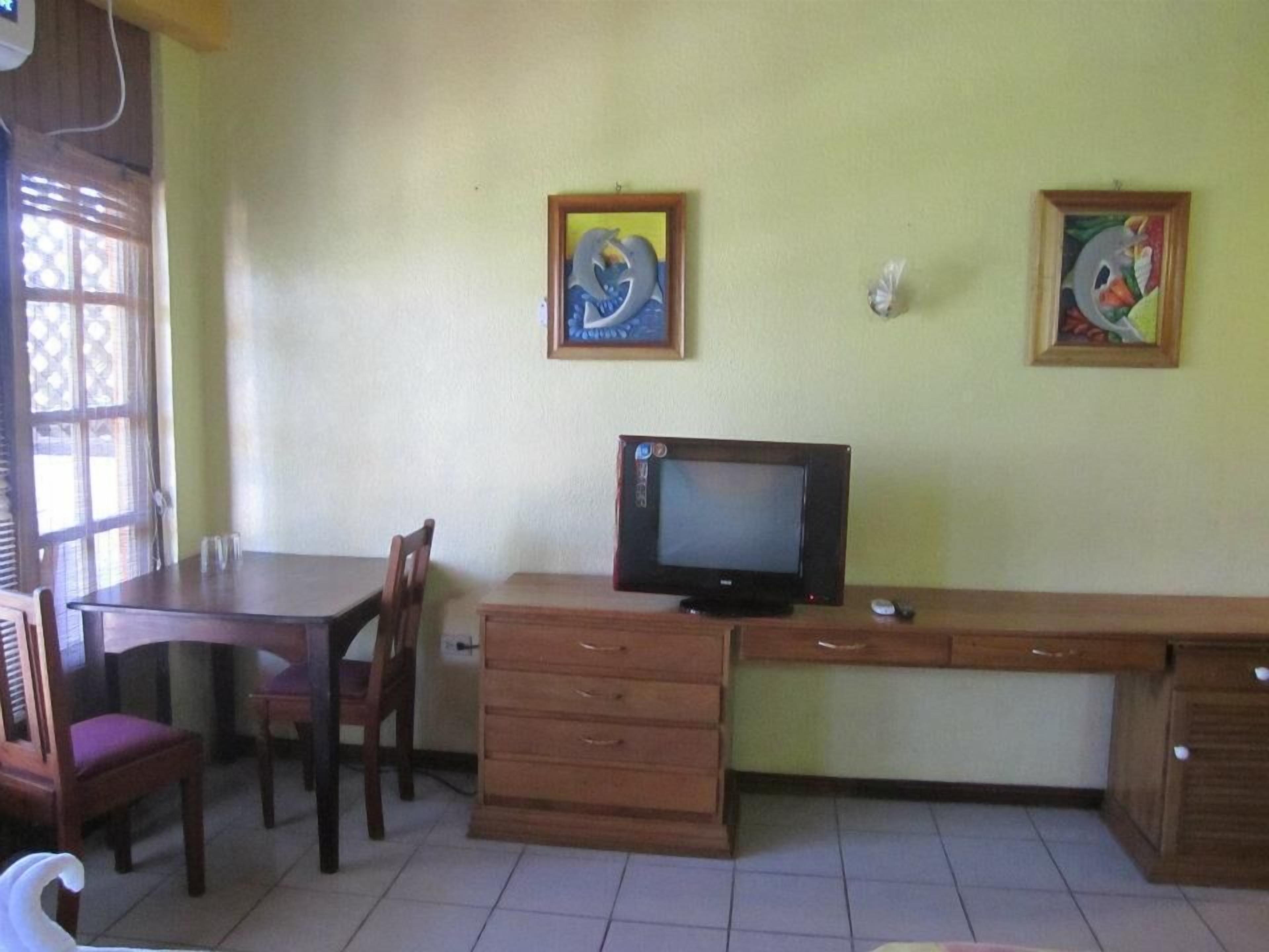 A room with a wooden desk, a television, two chairs, and two framed artworks on the wall.